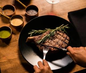 A person cutting into a large bone-in steak with rosemary