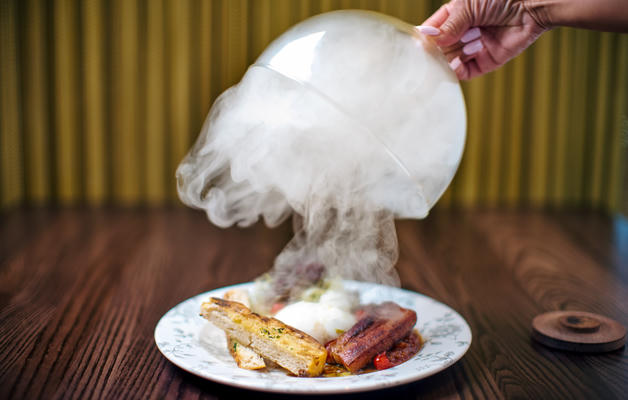A cloche being lifted by a gloved hand displaying artfully placed food and smoke
